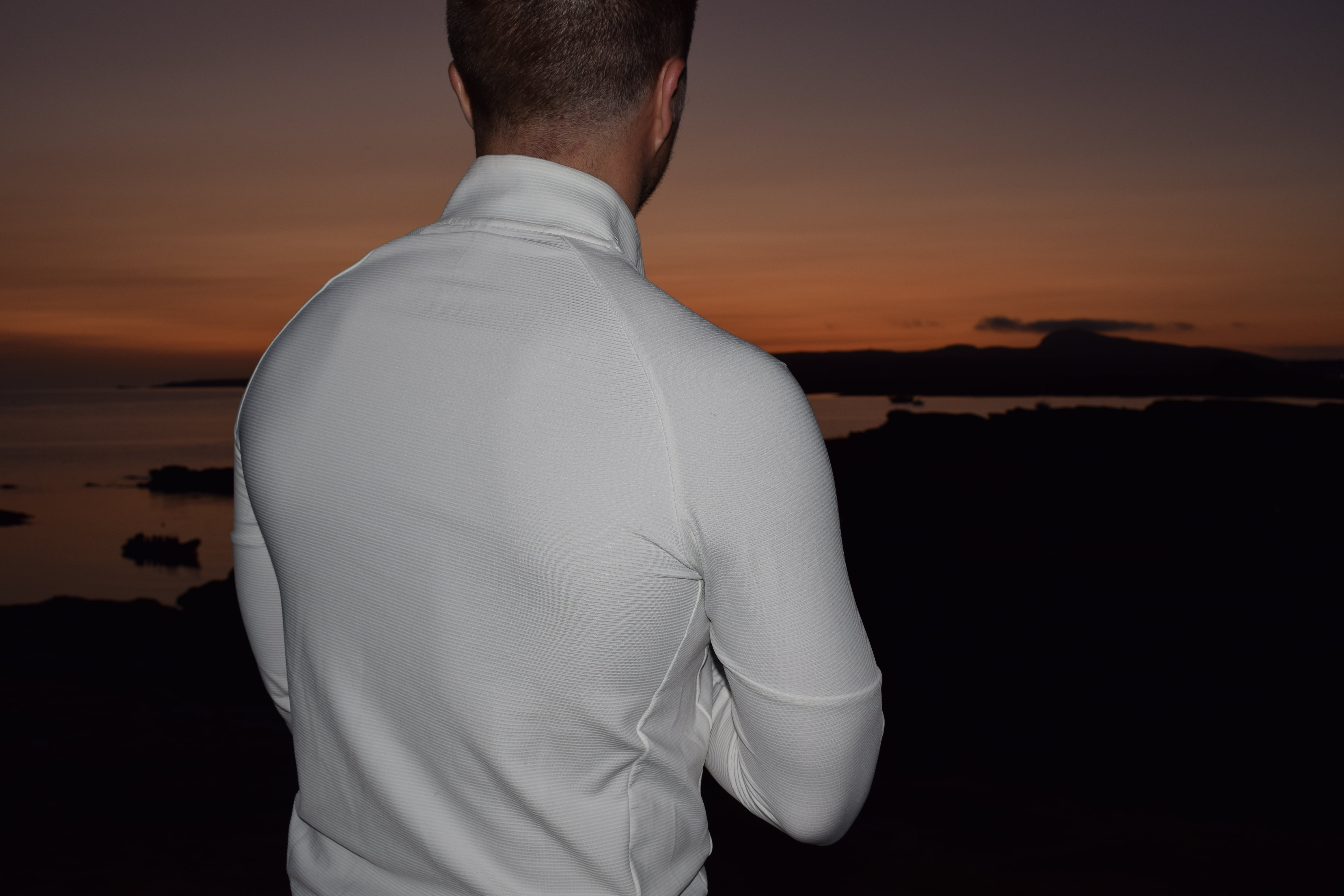 Man in a white half zip standing with his back to the camera, looking at a sunset over water